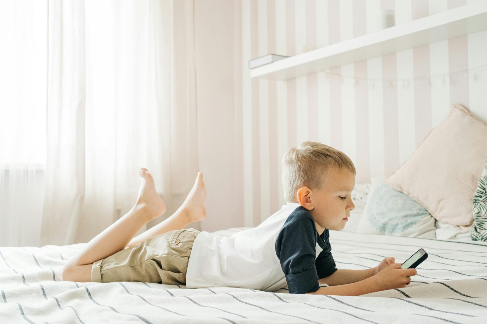 Young boy lying on bed using smartphone in a bright room, enjoying leisure time.