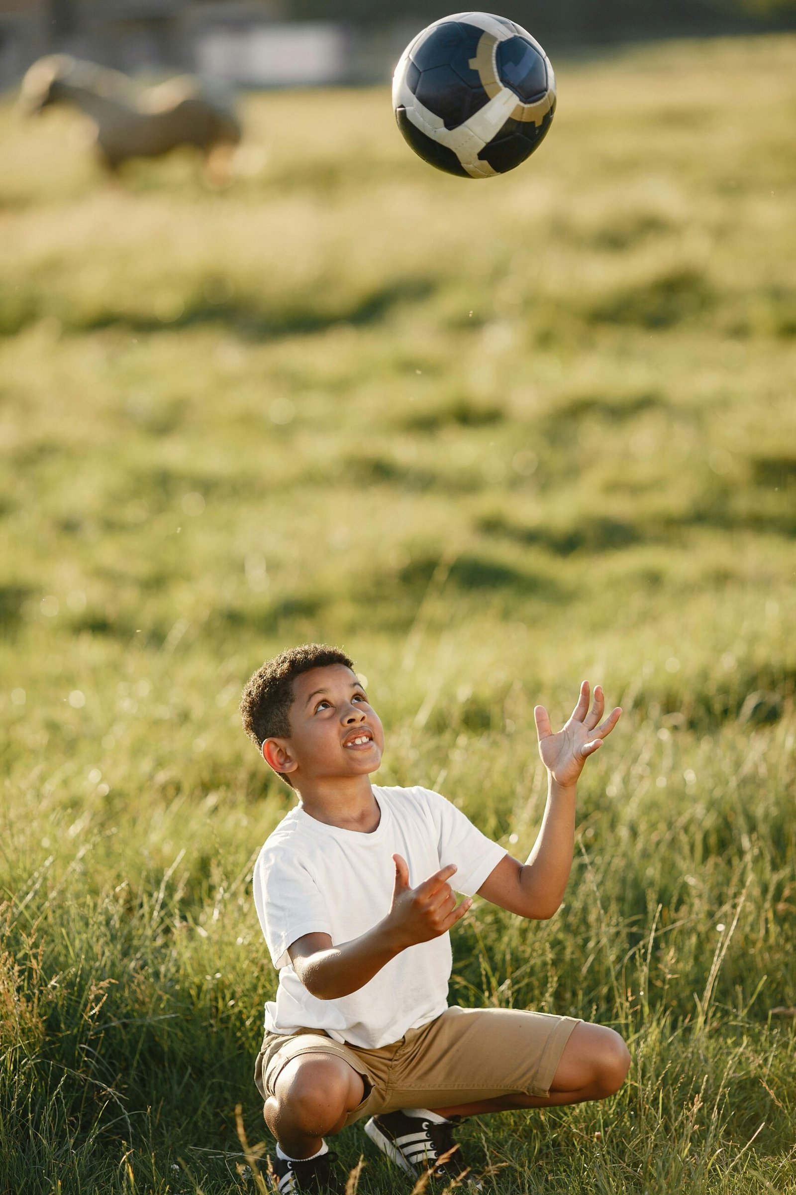 Child crouching in a grassy field, tossing a soccer ball into the air, in a warm outdoor setting.